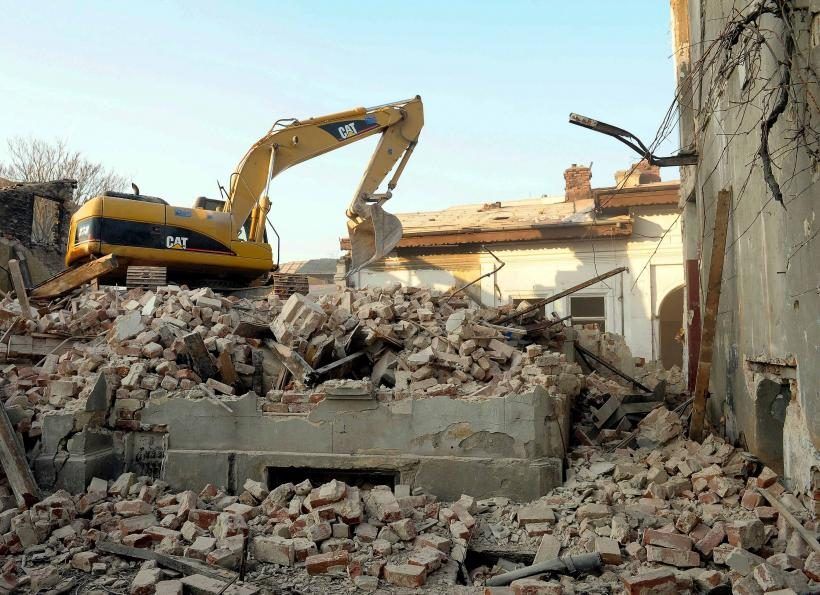 Excavadora CAT sobre escombros de ladrillo junto a un edificio antiguo parcialmente demolido y muros de sótano