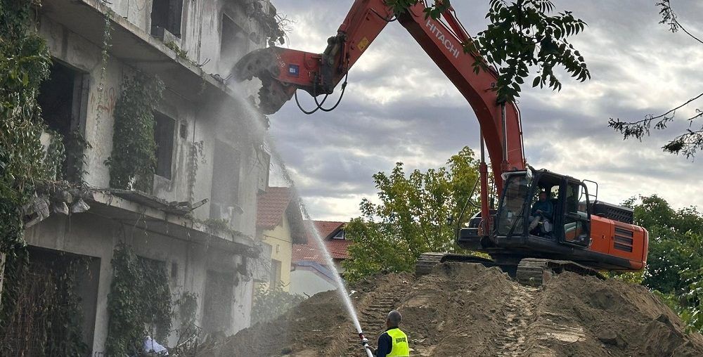 Hitachi excavator with a demolition crusher bringing down a multi-storey reinforced concrete building, with water spray for dust control