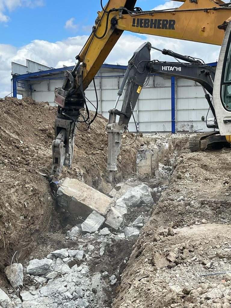 Breaking reinforced concrete ground beams with a Hitachi hydraulic breaker and a Liebherr excavator beside a steel hall